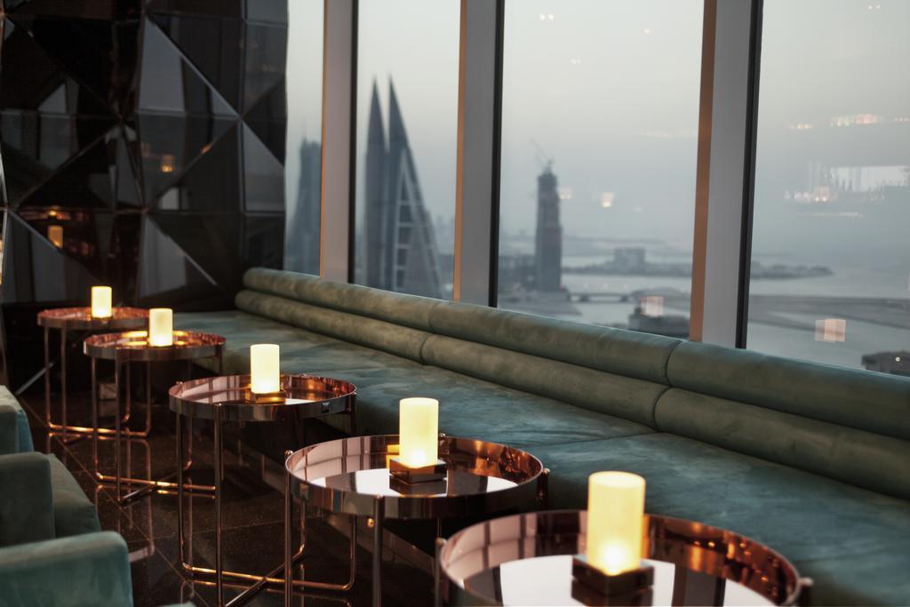 The Domain Bahrain Hotel And Spa - Adults Friendly 16 Years Plus Manama Exterior foto The photo depicts a stylish lounge area with a modern aesthetic. There are several circular tables made of metallic materials, each topped with a small glowing candle. A long, plush seating area in a teal color runs along the window, offering a view 